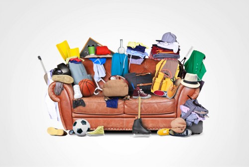 A brown upholstered sofa densely covered with a variety of discarded household and sports items, including clothing, shoes, toys such as a soccer ball and a basketball, a computer mouse, a rolled-up yoga mat, a yellow plastic container, a pair of sunglasses, and a beige handbag, with additional items like a small chalkboard, a washboard, and a blanket draped over the armrests. The background is plain white, emphasizing the cluttered appearance of the furniture. The scene reflects a mixture of domestic waste and personal belongings that might be gathered during rubbish removal services by Commercial Waste Brockley, serving the Brockley area in London. The assortment of materials and textures ranges from soft textiles and plastic containers to hard shoes and sports equipment, presenting an unorganized display of waste requiring professional collection. The environment appears to be a neutral session focused solely on the sofa, with no surrounding environment or premises visible, highlighting the need for waste clearance in urban living spaces.