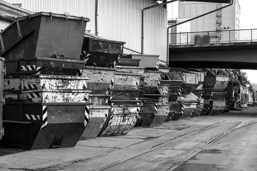 A row of large metal skips with a rectangular shape and reinforced sides, arranged along a concrete loading area adjacent to a warehouse or industrial building. The skips are used for waste collection and are visibly weathered, showing signs of rust and dirt on their surfaces. They are positioned on the left side of the image, with some stacked or slightly overlapping, indicating frequent use for rubbish removal services. In the background, a high metal railing is mounted on a platform attached to the building, and utility pipes or conduits run along the exterior wall, suggesting an industrial environment. The ground is concrete, with visible tire marks and slight grime, typical of a commercial waste management site. The lighting appears diffuse, consistent with an overcast day, emphasizing the utilitarian nature of the setting. This scene exemplifies the type of industrial waste skip storage managed by Commercial Waste Brockley in the Brockley area, supporting their rubbish and business waste removal services.