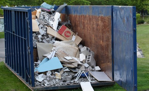 A large, industrial blue skip with partially rusted metal interior, filled with mixed waste materials including broken concrete slabs, various cardboard boxes, plastic debris, and scattered debris resting on the ground. The skip is situated outdoors on a grassy area with a paved surface nearby, and the background shows a well-maintained park or garden with green trees and grass, suggesting a typical waste collection site for commercial rubbish removal services in Brockley, London. The contents are piled unevenly with some materials spilling over the edges, indicating recent rubbish clearance from a commercial or domestic site. The scene is captured in natural daylight, highlighting the textures of the waste materials and the weathered appearance of the metal skip, aligning with professional waste management visual standards.