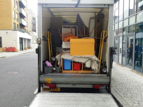 The image shows the open back of a grey commercial waste collection van parked on a residential street in Brockley, with the rear doors fully open to reveal a load of mixed household and office rubbish. Inside the van, there are several orange and black office chairs, some upright and others leaning, along with a yellow storage container. A small blue and red plastic storage box is positioned at the bottom, partially covered by a white shaggy rug. Various smaller items, including a black bag and a few scattered objects, are also visible among the rubbish. The setting features a modern residential area with multi-storey buildings on the left side and a glass-fronted commercial building on the right. The street is paved with grey concrete, and the van's license plate reads 'KUB1 LFV'. The scene suggests a business undertaking waste removal services in an urban environment, consistent with the offerings of Commercial Waste Brockley, targeting rubbish clearance in the Brockley area with a focus on domestic and small commercial waste disposal.