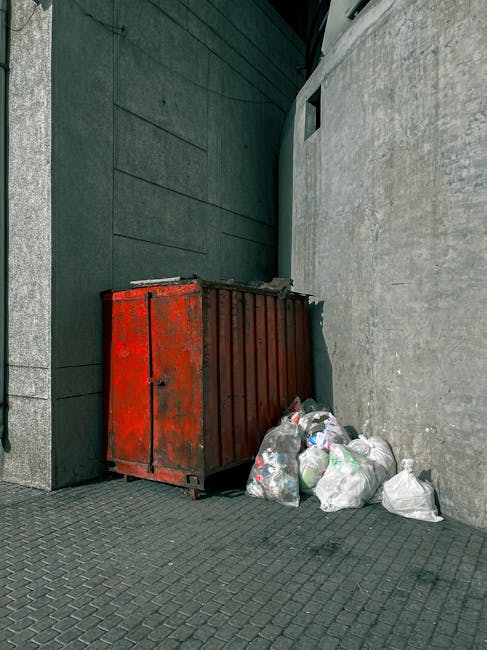 A narrow urban alleyway filled with overflowing rubbish and debris, including large black plastic bags, cardboard boxes, and miscellaneous waste items stacked against a weathered brick wall at the rea