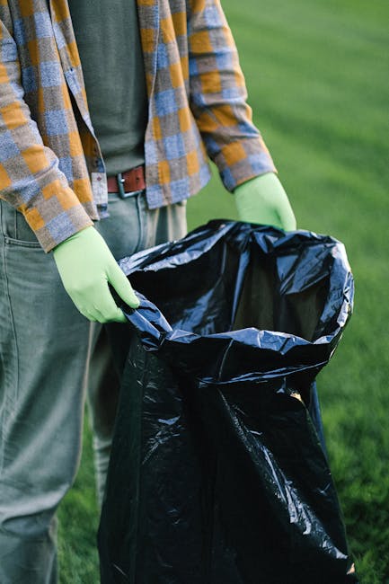 A person standing outdoors on a grassy area is holding a large black rubbish bag with their gloved hands. The individual is dressed in a checkered shirt featuring shades of yellow, grey, and blue, lay